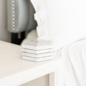 May include: A set of four white marble hexagonal coasters stacked on a white nightstand.