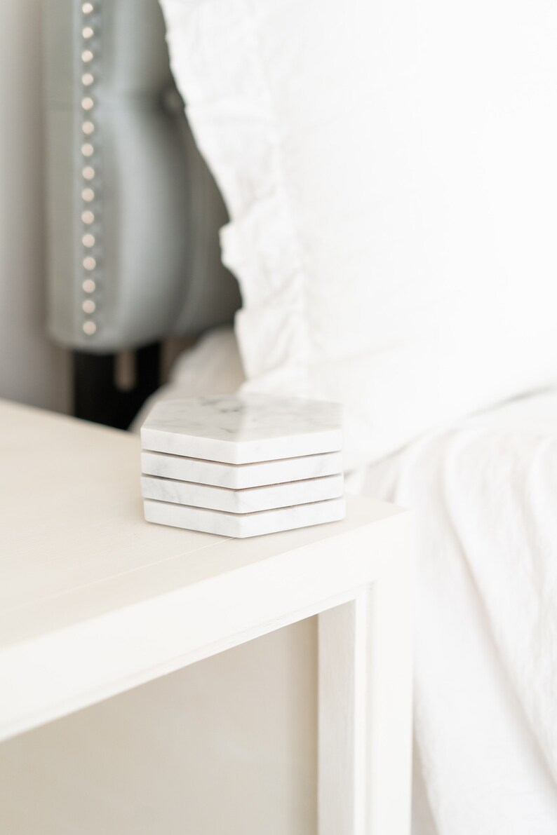 May include: A set of four white marble hexagonal coasters stacked on a white nightstand.