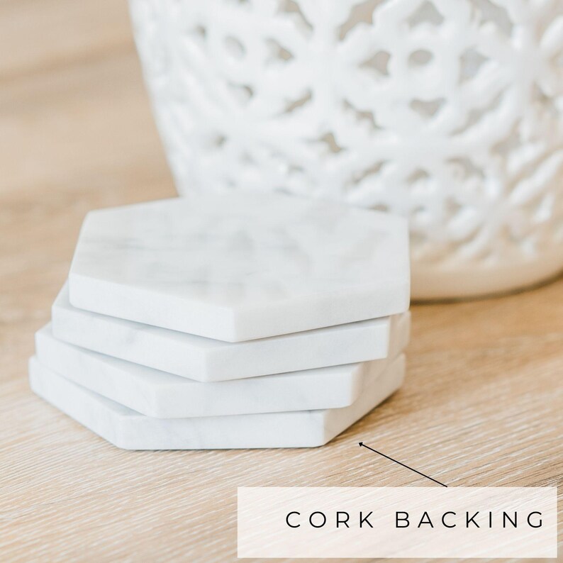 Stack of white Carrara marble hexagon coasters with soft grey veining, placed on a light wood surface. Text overlay with arrow reads “Cork Backing,” indicating the protective layer on the underside.