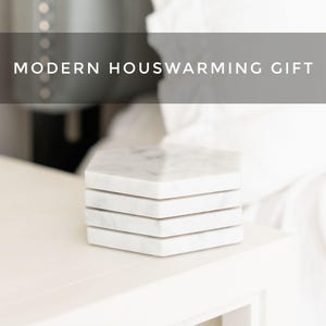 Elegant Carrara marble hexagon coasters with soft grey veining, neatly arranged on a light-toned bedside table. Text overlay reads “Modern Housewarming Gift.”