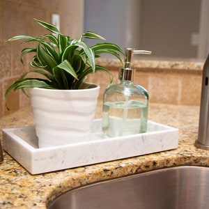 Carrara marble tray on a kitchen counter holding a potted plant and a glass soap dispenser. The tray's white surface with gray veining adds elegance to the sink area.