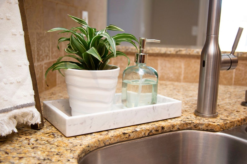 Carrara marble tray on a kitchen counter holding a potted plant and a glass soap dispenser. The tray's white surface with gray veining adds elegance to the sink area.