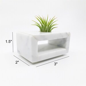 May include: White marble planter with a rectangular shape and a cutout in the middle. The planter is 3 inches wide, 2 inches deep, and 1.5 inches tall. A small air plant is sitting in the planter.