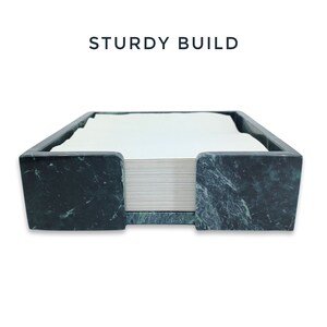 May include: A dark green marble napkin holder with a polished finish and a square shape, containing a stack of white paper napkins. The text STURDY BUILD is printed in dark blue letters above the holder, which features a front cutout.