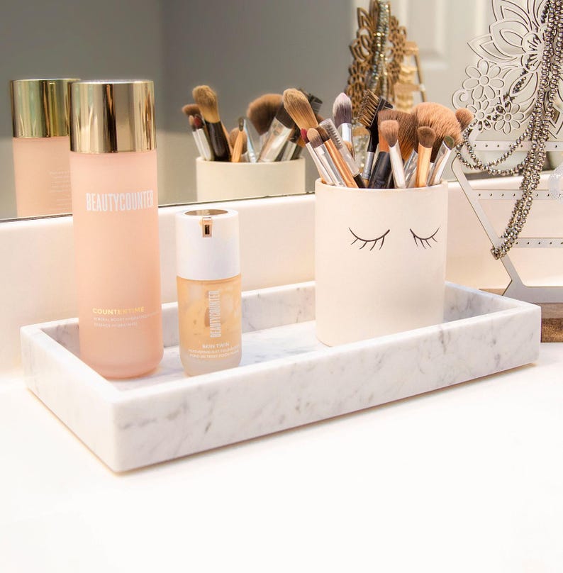 Carrara marble tray on a bathroom counter, holding beauty products and makeup brushes. Next to it is a jewelry stand displaying necklaces and earrings. The tray's white surface with gray veining adds a touch of elegance to the setup.