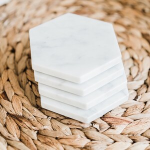 May include: A set of four white marble hexagonal coasters stacked on top of each other. The coasters are sitting on a woven brown wicker mat.
