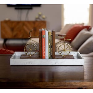 A rectangular Carrara marble tray is displayed on a wooden table, featuring a set of brass and wood bicycle bookends holding several books. The tray’s smooth surface and grey veining add a sophisticated touch to the living room setting.