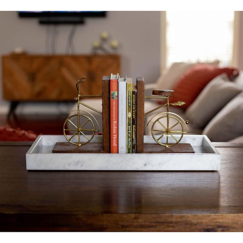 A rectangular Carrara marble tray is displayed on a wooden table, featuring a set of brass and wood bicycle bookends holding several books. The tray’s smooth surface and grey veining add a sophisticated touch to the living room setting.