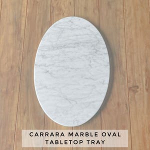 May include: Carrara marble oval tabletop tray. The tray is white with gray veining and has an oval shape. The text at the bottom of the image reads "CARRARA MARBLE OVAL TABLETOP TRAY."