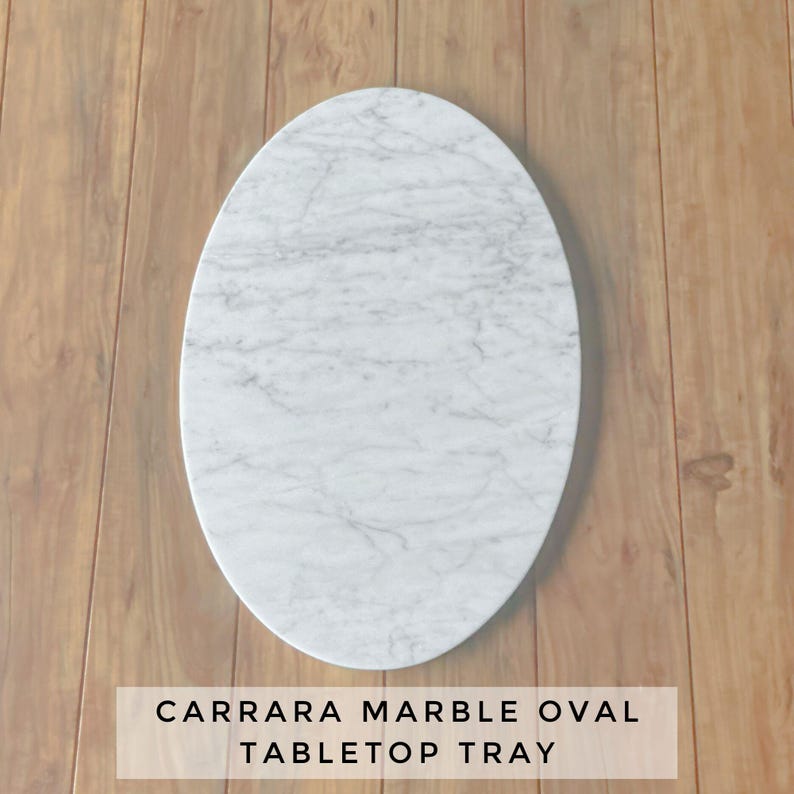 May include: Carrara marble oval tabletop tray. The tray is white with gray veining and has an oval shape. The text at the bottom of the image reads "CARRARA MARBLE OVAL TABLETOP TRAY."
