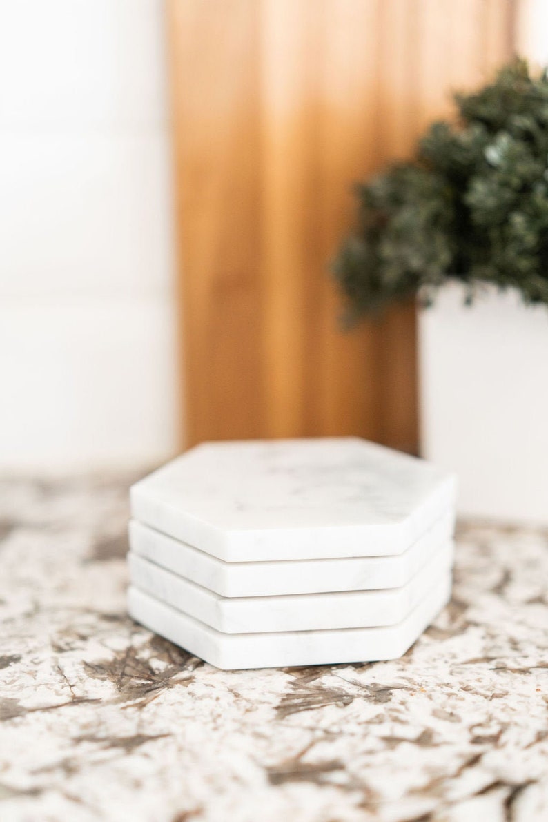 May include: A set of four white marble hexagon-shaped coasters. The coasters are stacked on top of each other and have a subtle marbled pattern.