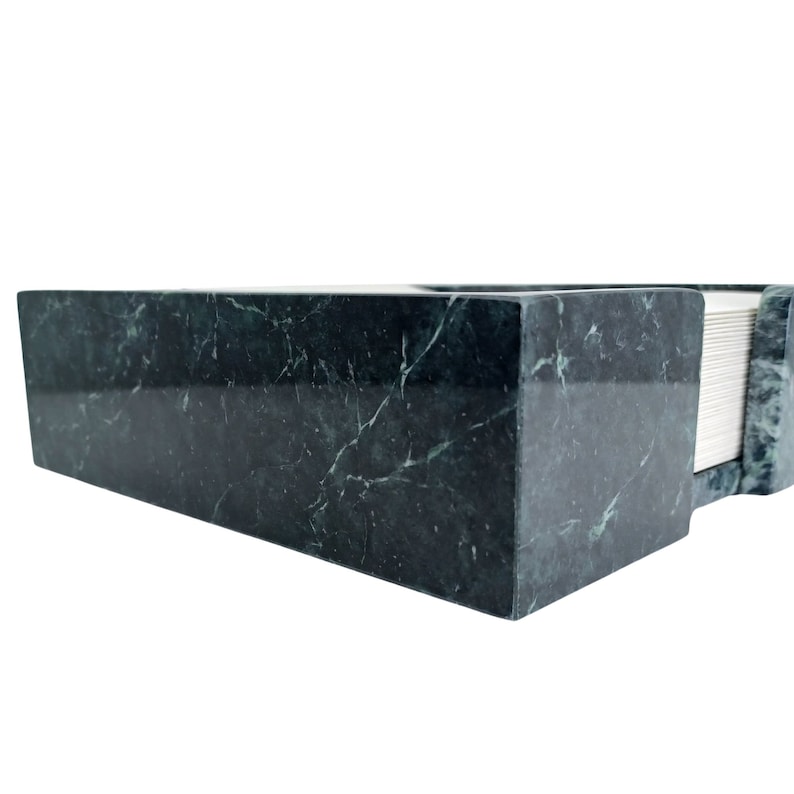 May include: A rectangular dark green marble napkin holder featuring a polished finish with natural white veining throughout the stone. The holder has a thick, solid construction and contains a stack of white paper napkins visible on the side.