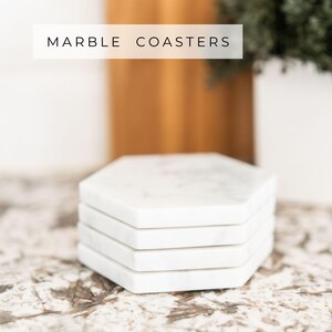 Stack of four handcrafted Carrara marble hexagon coasters with soft grey veining, shown on a marble surface. Text overlay reads “Marble Coasters.” Each piece features a cork backing to protect furniture.