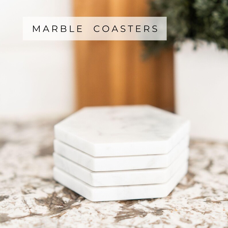 Stack of four handcrafted Carrara marble hexagon coasters with soft grey veining, shown on a marble surface. Text overlay reads “Marble Coasters.” Each piece features a cork backing to protect furniture.