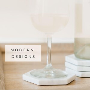 Glass of white wine resting on a white Carrara marble hexagon coaster with soft grey veining. Additional coasters stacked in the background. Text overlay reads “Modern Designs.”