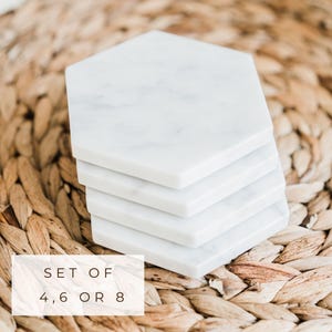 Stack of white Carrara marble hexagon coasters with subtle grey veining, displayed on a woven natural fiber placemat. Text overlay reads “Set of 4, 6 or 8."