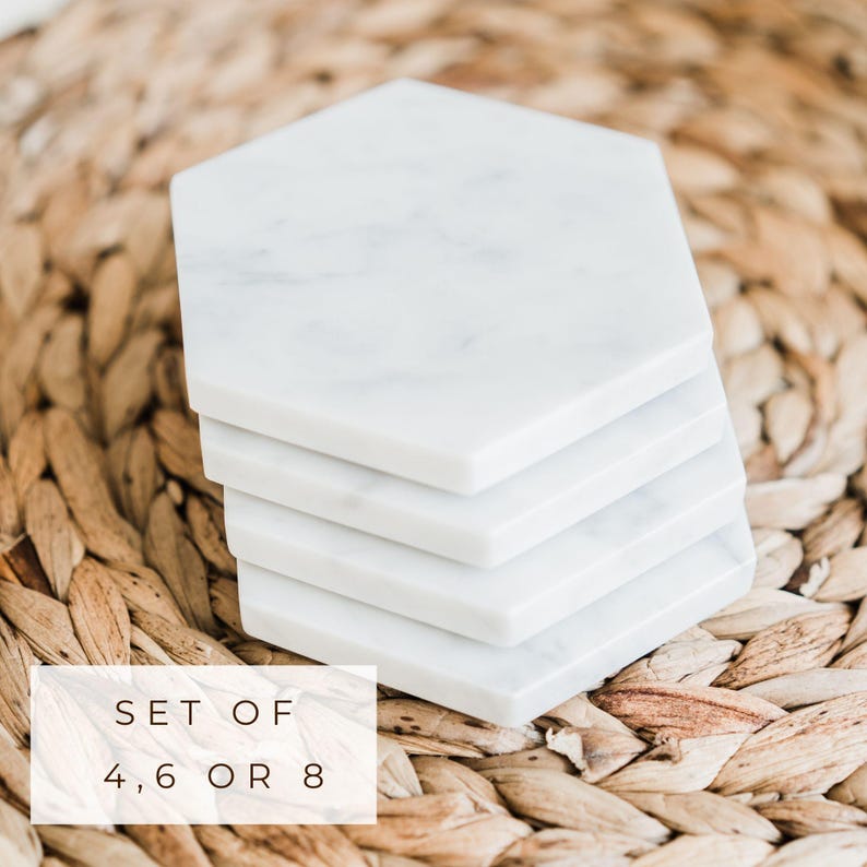 Stack of white Carrara marble hexagon coasters with subtle grey veining, displayed on a woven natural fiber placemat. Text overlay reads “Set of 4, 6 or 8."