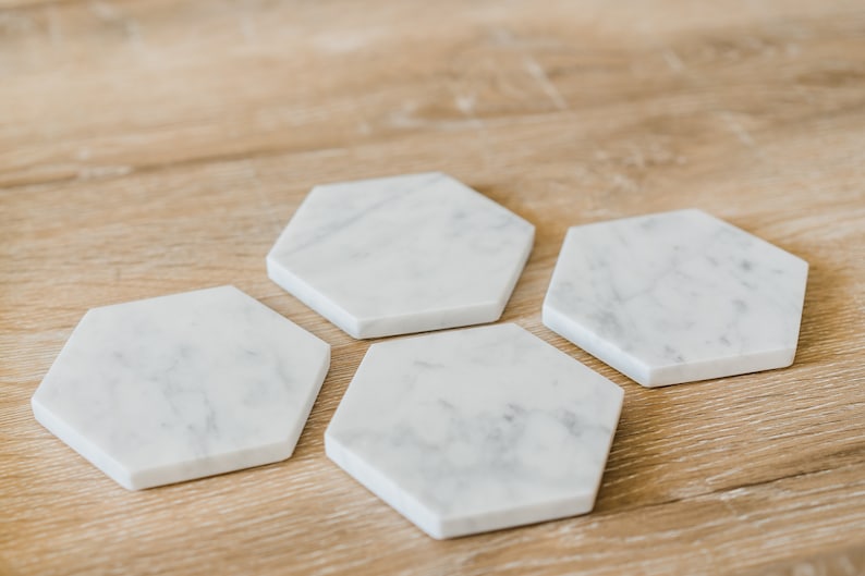 May include: Four white marble hexagon-shaped coasters on a light brown wooden surface.