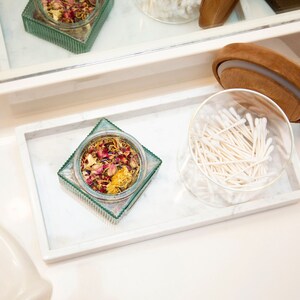 Carrara marble tray on a bathroom counter holding a glass jar with potpourri and a round container with cotton swabs. The tray's white surface with gray veining adds an elegant touch to the arrangement.
