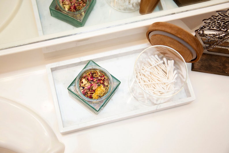 Carrara marble tray on a bathroom counter holding a glass jar with potpourri and a round container with cotton swabs. The tray's white surface with gray veining adds an elegant touch to the arrangement.