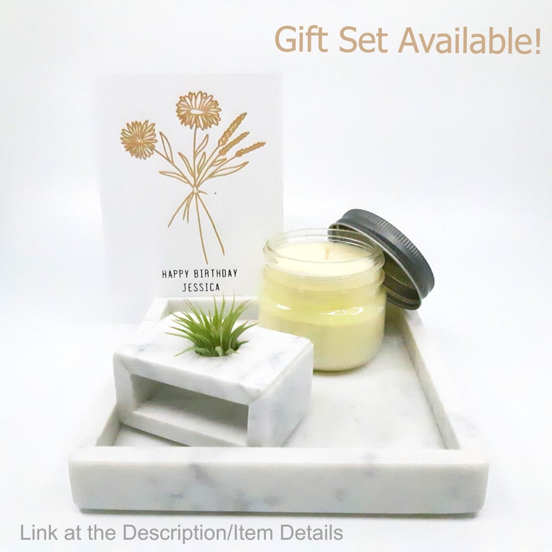 May include: A white marble tray with a white marble block, a small potted plant, a candle in a glass jar with a black lid, and a card with gold lettering that says "Happy Birthday Jessica" and a floral design.