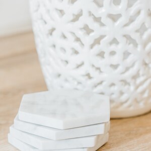 May include: A set of four white marble hexagonal coasters. The coasters are stacked on top of each other and are sitting on a light brown wooden surface.