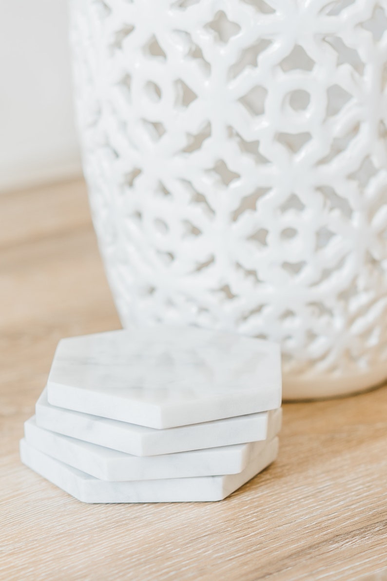 May include: A set of four white marble hexagonal coasters. The coasters are stacked on top of each other and are sitting on a light brown wooden surface.
