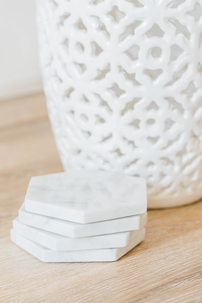 Set of handcrafted Carrara marble hexagon coasters with soft grey veining, displayed on a light wood surface next to a white ceramic vase. Elegant modern drink coasters with a polished or honed finish, available in sets of 4, 6, or 8.