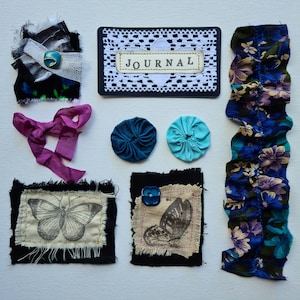May include: A selection of handmade fabric embellishments and journal elements. Features a journal cover with the word "JOURNAL", fabric flowers, butterfly prints, ribbons, and a ruffled fabric strip. Colours include black, blue, purple, and teal.