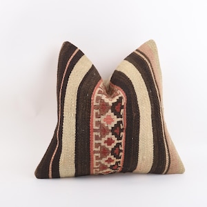Peut inclure: Un coussin décoratif avec un motif tissé. Il présente des rayures verticales marron, beige et rose. Le centre du coussin comporte une section à motifs avec des formes géométriques rouges, marron et roses. Le coussin est sur fond blanc.