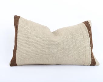 May include: A long, rectangular pillow with a beige woven fabric and brown trim. The pillow is lying flat on a white surface.