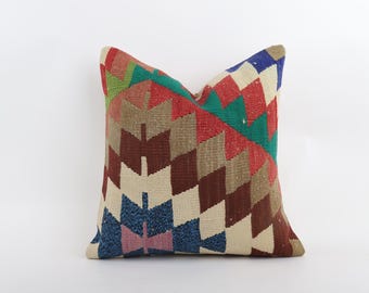 May include: Square decorative pillow with a geometric pattern in shades of red, green, blue, brown, and beige. The pillow has a woven texture and a symmetrical design, suitable for home decor.