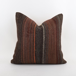 Puede incluir: Un cojín decorativo cuadrado con un patrón de rayas texturizadas en tonos marrón, naranja y gris. El cojín tiene un aspecto suave y acogedor, adecuado para añadir un toque de calidez a cualquier espacio habitable. El cojín mide aproximadamente 45 cm cuadrados.