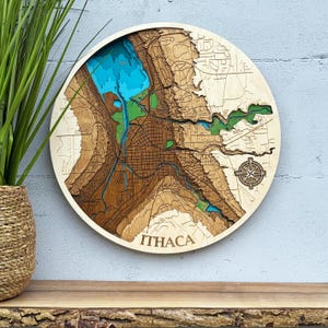 May include: A round, wooden map of Ithaca, New York, with layered topographic details. The map features blue water, green parks, and brown land, with the city name at the bottom. A compass rose is etched on the right side.