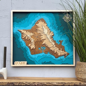 May include: A square wooden wall art piece featuring a layered 3D topographic map of the island of O'ahu. The map displays brown landmasses with raised mountain ranges surrounded by multiple shades of blue water. It includes a compass rose and the text O'AHU.