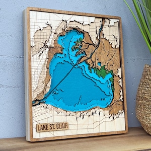 May include: A square wooden wall map of Lake St. Clair featuring layered, laser-cut topography. The water is depicted in bright blue, while the surrounding land areas are rendered in natural light wood tones with dark engraved grid lines.