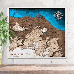 Puede incluir: Un mapa de madera en capas de Edimburgo, Escocia, con un marco blanco. El mapa presenta calles detalladas, puntos de referencia y contornos de elevación en tonos marrones y beige. El agua está representada en azul, con una rosa de los vientos en la esquina superior derecha.