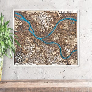 Peut inclure: Carte en bois de Pittsburgh avec une rivière bleue. La carte est encadrée de blanc et porte l'inscription "Pittsburgh" en haut et "November 9, 2019" en bas. Une rose des vents se trouve dans le coin inférieur gauche. Objet décoratif.