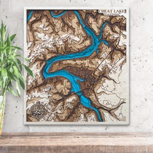 May include: Wooden topographic map of Cheat Lake, West Virginia, with a white frame. The map features a detailed layered design with brown and tan terrain, and a vibrant blue lake. A compass rose is in the lower left corner.
