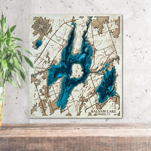 May include: A wooden map of Balsam Lake in Ontario, Canada. The lake is depicted in shades of blue, with the surrounding land in a light brown color. The map is framed in a light wood frame.