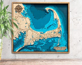 May include: A wooden 3D map of Cape Cod, Massachusetts, with a compass rose in the top left corner. The map is layered with shades of blue to represent the ocean and coastline. The text "CAPE COD Massachusetts" is printed in the bottom right corner.