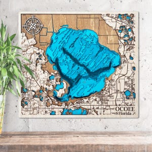 Può includere: Un'opera d'arte da parete in legno con una mappa dettagliata di Ocoee, Florida. La mappa evidenzia il lago con sezioni blu a strati, una rosa dei venti e i dettagli del territorio circostante. Il testo "OCOEE Florida" è visibile.