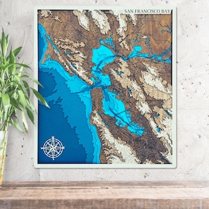 Pode incluir: Um mapa de madeira emoldurado da área da Baía de São Francisco. O mapa apresenta um design topográfico detalhado com tons de castanho, azul e branco. O texto "SAN FRANCISCO BAY" está no topo, e uma rosa dos ventos está na parte inferior.