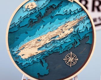 May include: A wooden map of Isle Royale National Park, featuring a detailed relief map of the island with a compass rose in the lower right corner. The map is painted in shades of blue and green, with a light brown wood finish for the island itself.
