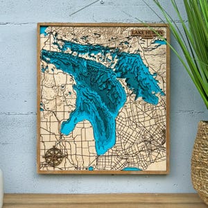 Può includere: Un'opera d'arte da parete quadrata in legno con una mappa batimetrica a strati del Lago Huron. La mappa mostra varie tonalità di blu per rappresentare la profondità dell'acqua, con masse terrestri in legno chiaro che mostrano strade. Una rosa dei venti.