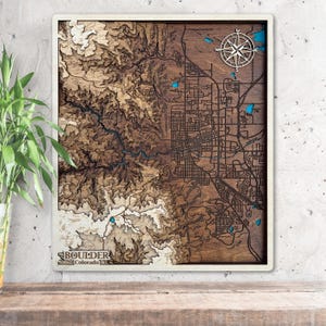Può includere: Mappa topografica in legno di Boulder, Colorado, con cornice bianca. La mappa presenta strati tagliati al laser che raffigurano la città e il terreno circostante, con una rosa dei venti e il nome della città in basso.