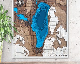Pode incluir: Uma peça de arte de parede em madeira com um mapa detalhado e em camadas do Lago Winnebago. O lago é retratado em tons de azul, contrastando com as áreas terrestres marrons e brancas. Uma rosa dos ventos está no canto superior esquerdo, e o nome do lago está na parte inferior.