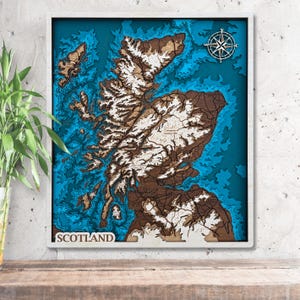 May include: A framed, multi-layered wooden map of Scotland, featuring shades of brown, white, and blue. The map includes a compass rose and the word "SCOTLAND" at the bottom. The artwork is set against a white frame.