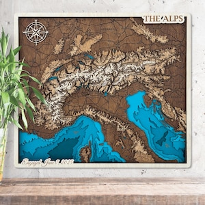 May include: A wooden map of the Alps, featuring a detailed topographical design with brown, white, and blue layers. The map includes a compass rose and the text "THE ALPS" and "Engaged June 7, 2025". The map is framed in white.