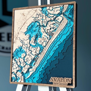 May include: A wooden map of Avalon, New Jersey, with a blue and teal color scheme. The map is raised and textured, showing the coastline and waterways. The words "Avalon, New Jersey" are printed in white on a black background at the bottom of the map.
